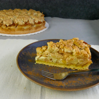 Im Hintergrund steht ein ganzer Streuselkuchen mit Apfel. Davor ein Stück Apfelkuchen auf einem Keramikteller mit einer Kuchengabel und Zimt bestreut.