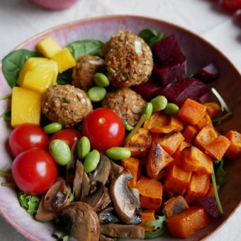 Auf dem Bild ist eine rosa Schüssel, die mit einer Bowl mit Buchweizenbällchen, Süßkartoffelwürfel, Tomaten, Edamame, Mango, Salat, Ruccola, Rote Bete und Pilzen gefüllt ist.