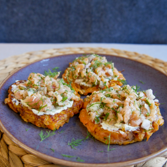 Auf einem weißen Tisch liegt ein Tischsetz aus Bast, darauf steht ein llila Teller und auf dem liegen drei süßkartoffelsösti mit Lachstatar und Dill