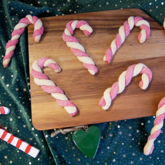 Auf einer dunkelgrünen Tischdecke mit kleinen goldenen Sternen liegt ein Holzbrett auf dem sechs Zuckerstangen Plätzchen verteilt sind. Eine Zuckerstange aus Holz liegt vorne links auf der Tischdecke. Daneben in der Mitte Vorne liegt ein dunkel grünes Herz aus Holz