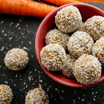 Auf einer schwarzen Schieferblatte steht eine kleine rote Schale mit Energyballs die in Kokos gewälzt wurden links neben der Schale liegen weitere drei Energyballs. Im Hintergrund links hinten liegen zwei Karotten.
