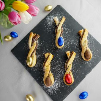 Auf einer grauen Tischdecke liegt eine schwarze Schieferntafel drumherum kleine goldenen und blaue Mini-Ostereier. Links hinten im Eck ist ein Tulpenstraus zu sehen. Auf der Schiederntafel liegen 5 Osterhäschen aus Bläterteig die mit Nutella gefüllt sind.