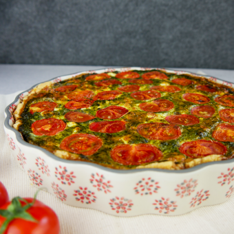 In einer weißen Quicheform mit roten gepunkteten Blumen außen ist eine gebackene Quiche mit Spinat und oben sind kleine Tomaten zu sehen