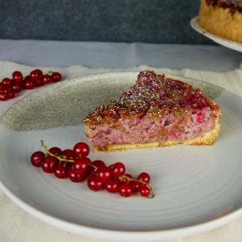 auf einem beigen Teller liegt im Vordergrund ein Strang Johannisbeeren dahinter ein Stück des Träubleskuchens. Hinter dem Teller ist rechts hinten ein drittel vom ganzen Kuchen zu sehen