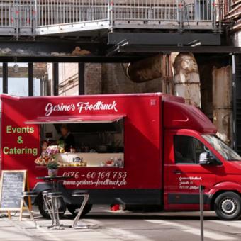 Roter großer Foodtruck mit der Aufschrift Gesines Foodtruck Davor steht ein Tich und 4 stühle und eine Tafel mit den Gerichten
