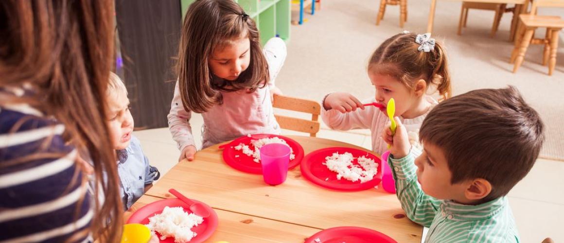 Kinder siten an einem gedeckten Tisch und erhalten Essen von der pädagoischen Fachkraft