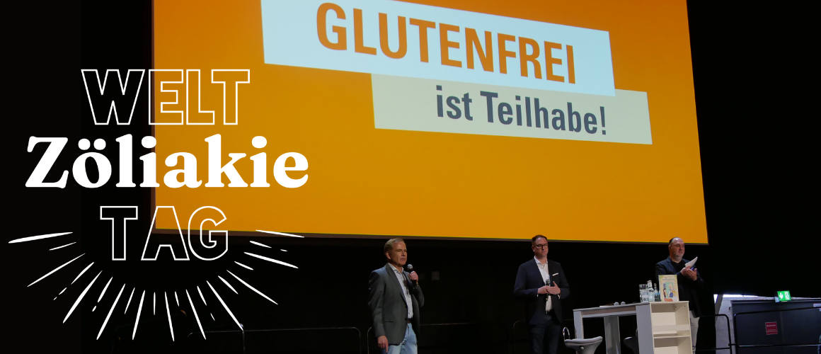 Auf einer Bühne stehen drei Personen vor einer großen orangefarbenen Leinwand. Auf der Leinwand steht der Slogan „GLUTENFREI ist Teilhabe!“. Links im Bild ist die Aufschrift „WELT Zöliakie TAG“ eingeblendet. Herr Gunnar Höckel spricht in ein Mikrofon, während die beiden anderen neben einem Stehtisch stehen. Die Aufnahme zeigt eine Veranstaltung der Deutschen Zöliakie Gesellschaft e.V. zum Thema Zöliakie und glutenfreie Ernährung.