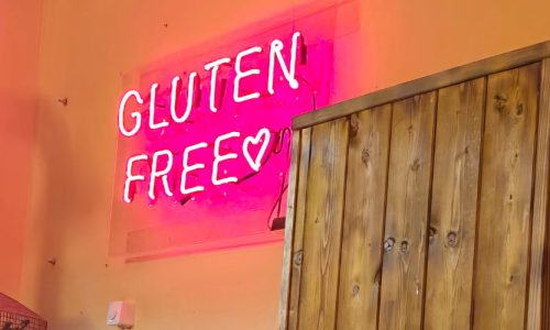 Pinkfarbene Leuchtschrift Glutenfree an einer ockerfarbenen Wand. Neben der Leuchtschrift steht ein robuster Holzschrank