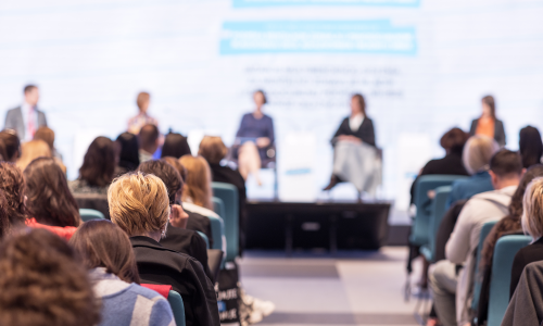 Zuschauer von Hinten vor Bühne auf der 5 Redner sitzen. Dahinter eine Präsentation