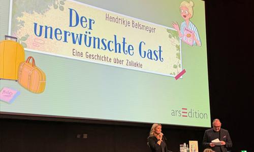 Auf einer Leinwand ist der Titel des Kinderbuches Der unerwünschte Gast eingeblendet. Am Pult sitzt Hendrikje Balsmeyer und liest aus ihrem Kinderbuch